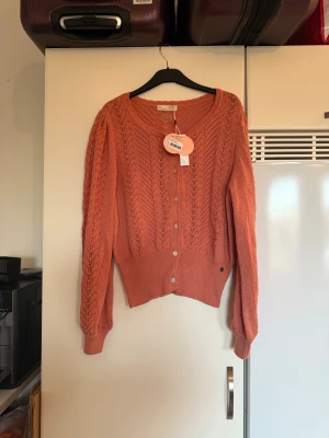 Rostfärgad kofta från Odd Molly - Snygg rostfärgad kofta från Odd Molly med v-ringning och dekorativa knappar framtill. Koftan är stickad i ett mönstrat material med hålstickade detaljer och har långa ärmar med breda muddar. Perfekt för dig som gillar unika och trendiga plagg. Storlek 4 som enligt deras storleksguide motsvarar en storlek L/XL. Helt ny med tags/ prislapp fortfarande kvar. 