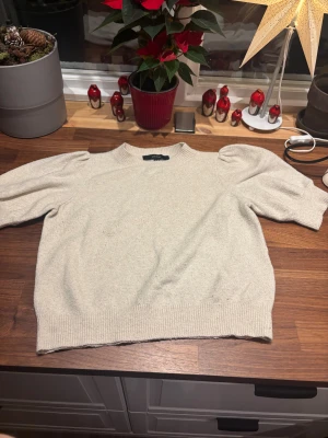 Beige stickad tröja Vero Moda S - Snygg beige stickad tröja från Vero Moda i storlek S. Tröjan har puffade korta ärmar och ribbade muddar vid hals, ärmslut och nederkant. Perfekt för dig som gillar enkel men trendig stil med en twist på axlarna.