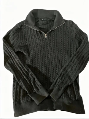 Mörkblå stickad half zip-tröja Riley - Mörkblå stickad tröja från Riley med kabelstickat mönster och half zip-dragkedja vid kragen. Tröjan har lång ärm och ribbade muddar vid ärmslut och nederkant. Perfekt för dig som gillar klassisk stil med en modern twist.