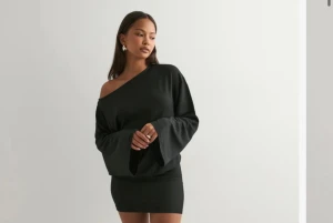 Svart oneshoulder kortklänning - Snygg svart kortklänning med oneshoulder-design och långa, vida ärmar. Klänningen har en tight passform nedtill och en lösare överdel som ger en cool kontrast. Perfekt för dig som gillar stilrena och trendiga plagg med lite edge.