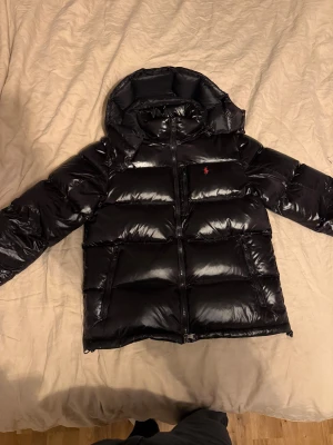 Polo Ralph Lauren The Gorham Shiny Down Jacket  - Säljer en svart, glansig dunjacka från Polo Ralph Lauren med huva och röd logga på bröstet. Jackan har två stora fickor med dragkedja, dubbla dragkedjor framtill och är fodrad för extra värme. Perfekt för kalla dagar och har en riktigt snygg streetstil. Riktigt puffig och håller än varm!