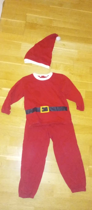 tomtedräkt / tomtepjamas 98/104 - Tomtedräkt i rött med vit kant och svart bälte med gul spänne tryckt på tröjan. Setet inkluderar matchande tomteluva med vit kant och boll. Mjuk bomullskänsla, perfekt för julens alla upptåg. Storlek 98/104.