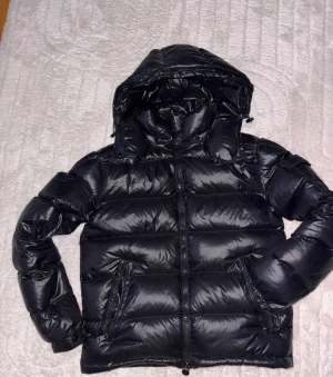 Svart Moncler pufferjacka med huva - Säljer en svart pufferjacka från Moncler med glansig finish och huva. Jackan har dragkedja framtill, två sidofickor med dragkedja och quiltad design. Insidan är grå och har en cool Moncler-etikett samt serietidningsdetalj. Perfekt för kalla dagar.