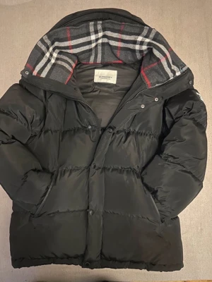 Svart pufferjacka Burberry M - Svart pufferjacka från Burberry i storlek M. Jackan har en klassisk Burberry-rutig huva i grått, vitt, svart och rött. Fylld med dun och har både dragkedja och knappar framtill. Perfekt för kalla dagar med sin tjocka och quiltade design.