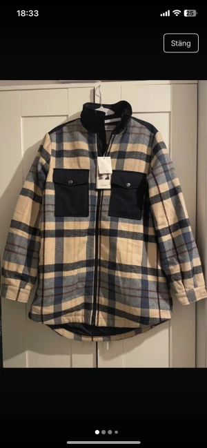 Rutig overshirt jacka från Fransa - Snygg overshirt jacka från Fransa med rutigt mönster i beige, blått och mörkblå detaljer. Jackan har två stora bröstfickor med knappar, dragkedja framtill och hög krage. Perfekt för lager-på-lager och chill stil.