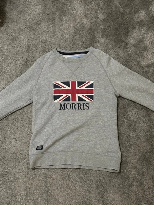 Grå sweatshirt från Morris  - Grå sweatshirt från Morris i bomull med storbritaniens flagga och Morris-tryck på bröstet. Klassisk rund halsringning och långa ärmar. Perfekt för dig som gillar stilren design med brittisk touch. Det är storleken S men skulle säga att den passar mer som en M. Hör av er vid frågor och funderingar.