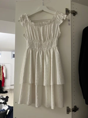 Vit kort volangklänning med prickar - Superfin vit kort klänning med volanger och små prickar i tyget. Klänningen har offshoulder-ärmar med rynkade detaljer och smock i midjan för en snygg passform. Perfekt för sommarens alla tillfällen, lätt och luftig med romantisk vibe.