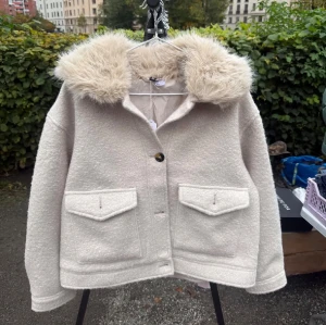 Beige jacka med fuskpäls-krage - Snygg beige jacka med stor fluffig fuskpäls-krage och två stora fickor framtill. Jackan har en rak passform, stängs med knappar och har en mjuk, ullig känsla. Perfekt för kyliga dagar när du vill vara både varm och stilren.