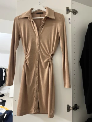 Beige kortklänning med kedjedetalj - Snygg beige kortklänning från NA-KD med mjukt stretchigt material och lång ärm. Klänningen har knappar hela vägen framtill, markerad midja med cool guldfärgad kedjedetalj på sidan och klassisk krage. Perfekt för dig som gillar trendiga detaljer och stilren look.