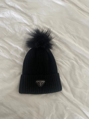 Svart ribbstickad mössa  - Säljer en svart ribbstickad mössa med stor fluffig pälsboll på toppen och ikonisk metallogo framtill. Mössan har bred uppvikt kant och är riktigt snygg till vinterlooken. Perfekt accessoar för dig som vill ha både stil och värme.