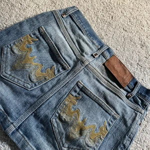 Jeanskjol  - Säljer en klassisk blå jeanskjol med snygga guldiga broderier på bakfickorna. Kjolen är kort, har fickor och stängs med knapp och dragkedja. 