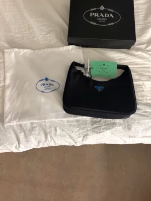 Svart axelväska från Prada - Snygg svart axelväska från Prada med klassisk, stilren form och ikonisk trekantig logga framtill. Väskan är gjord i syntetmaterial och har en enkel design med dragkedja upptill. Perfekt storlek för det viktigaste och kommer med originalförpackning och dustbag.