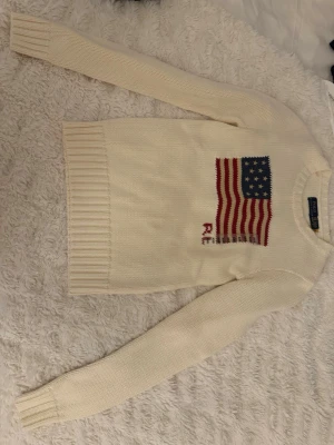 Vit stickad tröja från Ralph Lauren - Snygg vit stickad tröja från Polo Ralph Lauren med amerikansk flagga i blått och rött på bröstet. Storlek XXS. Aldrig använd. Skickas nytvättad. Nypris: 4895kr