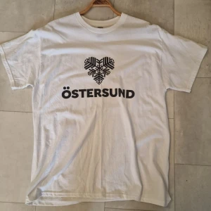 Vit Östersund t-shirt med tryck - Vit t-shirt med svart tryck framtill där det står Östersund och ett stiliserat hjärta i snöflinge-design. Klassisk rund hals och korta ärmar. Perfekt för dig som vill visa kärlek till staden med en enkel och clean look. Bomull.