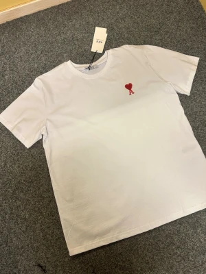 Vit t-shirt från Ami med rött hjärta - Vit t-shirt från Ami med klassisk rund hals och korta ärmar. På bröstet finns ett broderat rött hjärta med bokstaven A. T-shirten är gjord i mjuk bomull och har en clean, stilren look som är lätt att matcha med allt.