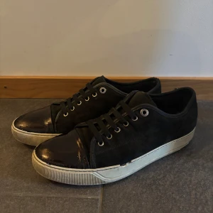 Lanvin  - Snygga svarta sneakers från Lanvin med ovandel i mocka och en glansig, lackad tå. Skorna har vita gummisulor och svarta skosnören. Metallfärgade öljetter ger en clean look. Perfekta för dig som gillar stilrena och exklusiva sneakers. Sulan är lite gul 