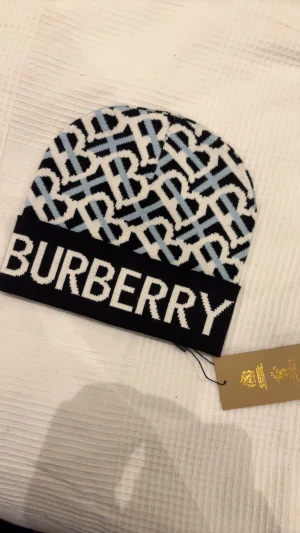 Burberry mönstrad mössa i blått och vitt - Snygg mössa från Burberry med stort logotryck och ett grafiskt mönster i ljusblått, vitt och svart. Mössan är stickad och har en mjuk känsla, perfekt för att lyfta din vinterstil. Ikonisk design med Burberrys signaturmönster och bred kant.