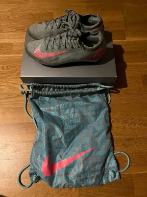  Nike fotbollsskor  - Säljer ett par grå Nike fotbollsskor med rosa swoosh och coola detaljer i blått på hälen. Skorna har snörning och är tillverkade i ett syntetmaterial som ger bra känsla på planen. Medföljer en matchande Nike-påse i grått och turkos. Perfekt för dig som vill sticka ut på fotbollsplanen. Liten skavank på höger fot (visas på bilden). Skobox oxh skopåse medföljer!