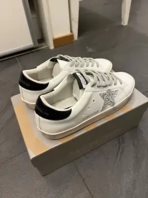 Golden Goose sneakers i vitt skinn med svart häl och silverglittrig stjärna på sidan. Klassisk låg modell med grå snörning och platt sula. Snygg GGDB-detalj på hälen och tungan. Kommer med originalkartong och påse.