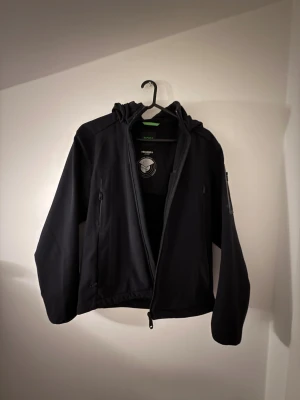 Svart vindjacka från BOSS med huva original pris 6500 - Helt ny, använt max 1 vecka. Original priset var 6500kr Svart vindjacka från BOSS med huva och dragkedja framtill. Jackan har diskreta logodetaljer på ärmen, flera praktiska fickor med dragkedja och är tillverkad i ett slätt syntetmaterial. Perfekt för blåsiga dagar och enkel att matcha med allt. 