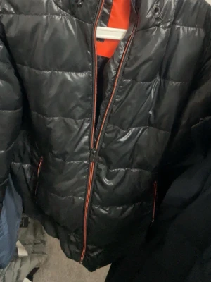 Svart pufferjacka från Björn Borg - Svart pufferjacka från Björn Borg Sport med orange detaljer och dragkedja. Jackan har quiltad design, lång ärm och insida med prickigt mönster och orange foder. Perfekt för kalla dagar när du vill ha både stil och värme.