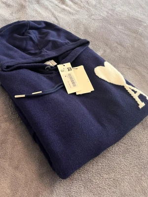 Ami Paris tröja  - Säljer en mörkblå hoodie från Ami Paris med ikoniskt hjärta och A på bröstet. Tröjan har huva med dragsko, känguruficka och ribbade muddar. Tillverkad i mjuk ull för extra komfort och stilren look.