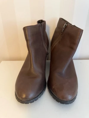 Bruna stövletter med kilklack från XIT - Säljer ett par bruna stövletter från XIT i storlek 38. Skorna har kilklack och är tillverkade i skinn med dragkedja på sidan. Snygg rund tå och stabil klack i trälook. Perfekt för dig som gillar stilrena boots med lite höjd.