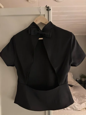Svart bolerotopp med rosett - Säljer en svart topp med korta ärmar och hög krage. Toppen har en snygg rosettdetalj baktill med öppen rygg. Aldrig använd! Säljer då den inte kom till användning.