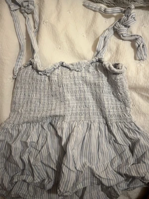 Blåvit randig kortklänning från Nelly - Supersöt blåvit top från Nelly i storlek XXS. Toppen  har smock upptill, volangig kjol och knytbara axelband. Tunt och luftigt material med smala ränder, perfekt för varma dagar. Ger en fräsch och somrig vibe!