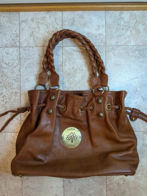 Brun handväska från Mulberry i skinn - Säljer en snygg brun handväska från Mulberry i äkta skinn med flätade handtag och detaljer i mässing. Väskan har snörning upptill och en stor Mulberry-logga framtill. Insidan har dragkedja med Mulberry-träd på läderfliken. Perfekt storlek för allt du behöver.      Inte äkta .