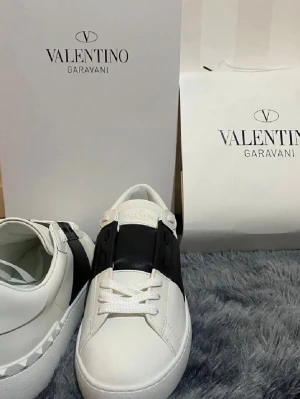 Valentino Garavani skor  - Säljer ett par snygga vita Valentino Garavani sneakers med svart detalj över vristen och coola nitar på hälen. Skorna har rund tå, platt sula och är gjorda i skinn. Perfekta för dig som vill ha en stilren och lyxig look.