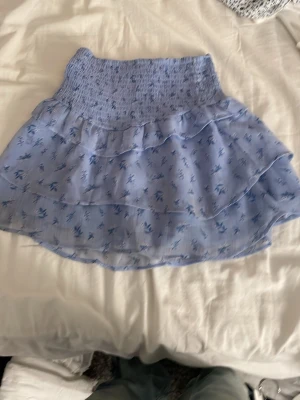 Blommig ljusblå volangkjol - Supersöt ljusblå kortkjol med små blå blommor och volanger. Kjolen har en smockad midja som gör den bekväm och sitter snyggt. Perfekt för dig som gillar romantisk och feminin stil. Materialet känns lätt och luftigt.