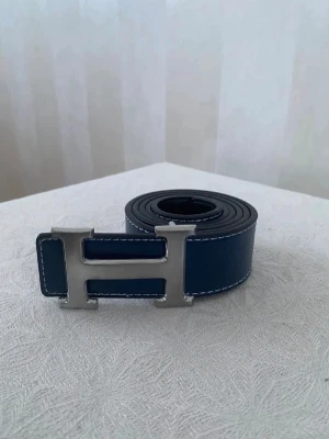 Mörkblå skinnbälte med H-spänne - Snyggt mörkblått bälte med ett stort H-format metallspänne. Bältet är tillverkat i skinn med vita kontrastsömmar längs kanterna som ger en cool detalj. Perfekt accessoar för att lyfta din outfit med en stilren touch.