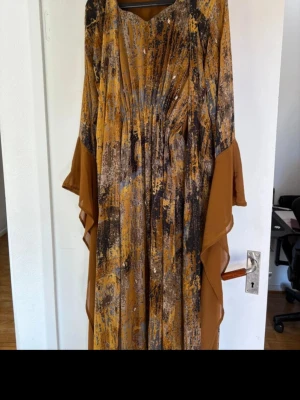 kurdisk klänning - Säljer en unik långklänning med abstrakt mönster i bruna, gula och grå toner. Klänningen har glittriga detaljer och pärlor på framsidan samt vida ärmar med volang. Materialet känns lätt och luftigt, perfekt för dig som gillar att sticka ut med din stil.
