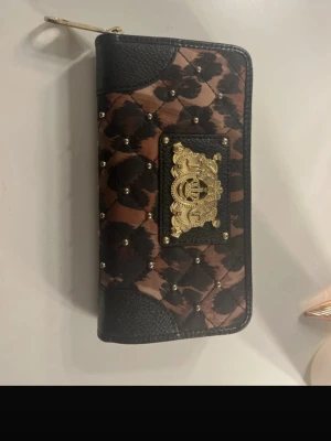 Leopardmönstrad plånbok Juicy Couture - Snygg plånbok från Juicy Couture med leopardmönster i brunt och svart, quiltad design med små guldfärgade nitar och ett stort guldfärgat emblem framtill. Plånboken har flera fack, dragkedja runtom och detaljer i svart skinnimitation. Perfekt för dig som gillar statement-accessoarer. Ena drag kedja grejen man drar i har tappads som man ser på bilden har lite skavanker. Köpte på en vintage affär för 1099kr