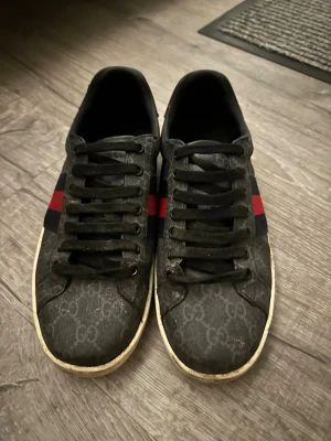 Svarta Gucci sneakers med röd/blå rand - Box medföljs fråga för bild på den om du är intresserad ✅✅!!!ALLT SMUTS KAN TAS BORT KOSTAR EXTRA OM DU VILL ATT JAG SKA TVÄTTA DE. PRIS EJ HUGGET I STEN✅✅!!’Säljer ett par svarta Gucci sneakers med klassiskt GG-mönster och röd/blå rand på sidan. Skorna har svarta snören, vit sula och rund tå. Materialet är skinn med tryckt monogram och detaljer i textil. Perfekta för dig som vill ha en lyxig touch till din outfit.
