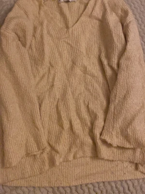 Beige stickad tröja från Mango - Mysig beige stickad tröja från Mango med v-ringad hals och långärmad design. Tröjan har en ribbad struktur som ger en cool och avslappnad look. Perfekt för lager-på-lager under hösten och vintern. Passar dig som gillar enkel och stilren stil.