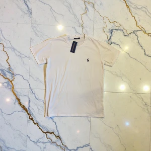 Vit t-shirt från Polo Ralph Lauren - Säljer en stilren vit t-shirt från Polo Ralph Lauren med klassisk rund hals och korta ärmar. T-shirten har en liten broderad logga i svart på bröstet och är gjord i mjuk bomull som känns skön mot huden. Perfekt för en clean och enkel look.