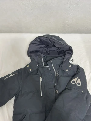 Svart dunjacka från Moose Knuckles - Säljer en svart dunjacka från Moose Knuckles med stor huva, silverfärgade dragkedjor och snygga metallknappar. Jackan har ett coolt logomärke på ärmen och flera praktiska fickor. Perfekt för kalla dagar och riktigt stilren look.