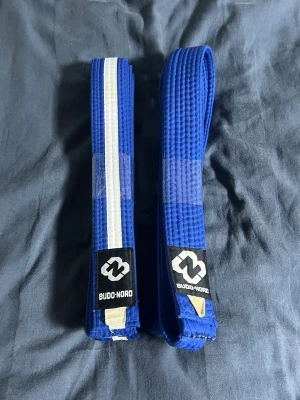 Blåa karate bälten - Bilderna visar två blåa bälten från Budo-Nord, inte några skor. Bältena är av bomull och har etiketter med storleksinformation. Ett av bältena har en vit rand längs hela längden.