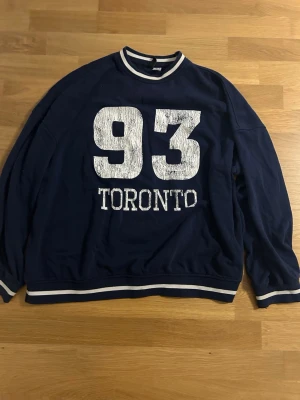 Mörkblå sweatshirt - Säljer en mörkblå sweatshirt från h&m i storlek M. Tröjan har vita ränder vid krage, ärmslut och nederkant samt ett stort tryck med siffran 93 och texten TORONTO på framsidan. Materialet är mjuk bomull och modellen är normal passform med långärmade ärmar.