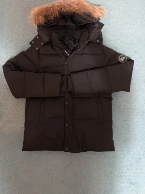 Svart dunjacka från Canada Goose M - Svart dunjacka från Canada Goose med stor huva och fluffig pälsdetalj. Jackan har flera fickor, knappar och dragkedja framtill samt ett snyggt märke på ärmen. Perfekt för kalla dagar och har en riktigt schysst oversized look.