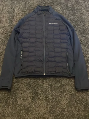 Mörkblå quiltad jacka Peak Performance - Snygg mörkblå jacka från Peak Performance i storlek L. Jackan har quiltad framsida och rygg, hel dragkedja och två sidofickor med dragkedja. Materialet är polyester och jackan har en sportig look med långärmade ärmar.