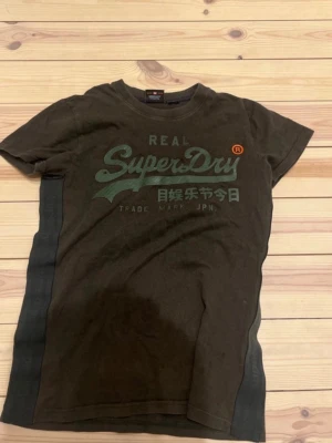 Mörkgrön Superdry t-shirt med tryck - Säljer en mörkgrön t-shirt från Superdry med stort grönt logotryck och japanska tecken på bröstet. T-shirten har korta ärmar och rund hals. Sidorna har diskreta band med Superdry-text. Perfekt för dig som gillar streetwear och snygga detaljer. Hör av er vid frågor mvh Nils i storlek S 