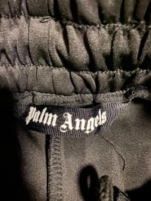Palm Angels svarta mjukisbyxor med ränder - Svarta mjukisbyxor från Palm Angels med vita sidorevor och logga broderad på vänster ben. Byxorna har resår i midjan med snörning och raka ben. Tillverkade i ett glansigt syntetmaterial som ger en sportig vibe. Perfekta för en streetwear-look.