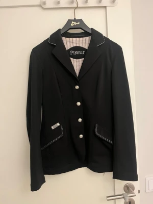 Svart ridkavaj från Pikeur - Säljer en svart ridkavaj från Pikeur med snygga silverfärgade knappar och vit kontrastsöm vid kragen och fickorna. Kavajen har klassisk passform, lång ärm och är tillverkad i ett slätt syntetmaterial. Perfekt för dig som vill ha en stilren look på tävling eller träning. Pris går att diskutera🥰🥰