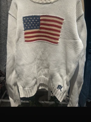 Vit stickad tröja Polo Ralph Lauren - Vit stickad tröja från Polo Ralph Lauren med amerikansk flagga i blått, rött och beige på bröstet. RL-broderi i blått vid nederkanten. Tröjan har rund hals, långa ärmar och ribbade muddar. Perfekt för dig som gillar klassisk och preppy stil.