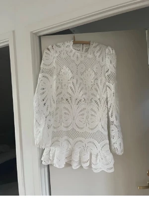 Vit spetsklänning med volang - Superfin vit kort klänning i spets med långärmade puffärmar och volangkant nertill. Klänningen har ett detaljerat broderat mönster, hög hals och dragkedja i ryggen. Perfekt för dig som vill sticka ut med en romantisk och trendig look.