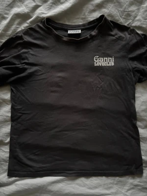 Grå Ganni Loveclub t-shirt - Grå t-shirt från Ganni med vit 'Ganni Loveclub'-print fram och stor 'LOVECLUB'-text på ryggen. Klassisk rund hals och korta ärmar. Mjuk bomullskvalitet som känns chill och enkel att styla till jeans eller shorts.