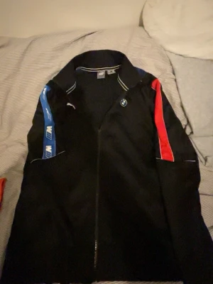 Svart BMW Motorsport jacka från Puma - BMW Motorsport jacka från Puma i svart med blå och röd detalj på ärmarna. Jackan har dragkedja framtill, vit piping och broderad BMW-logga på bröstet. Materialet är polyester och passformen är normal. Perfekt för dig som gillar motorsport och snygg streetwear.